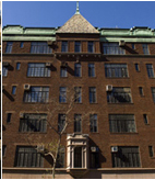 The apartment building is built in red bricks. The main door or entrance is at the center. The upper top or the rooftop is painted in old minty green color. An old tree barely no leaves is facing the entire apartment.